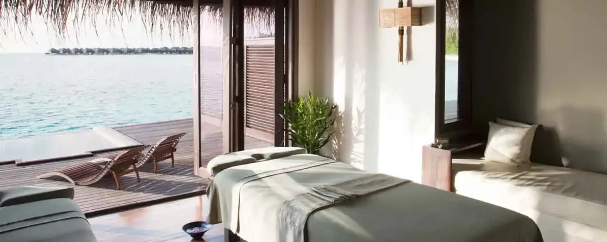 Coco Bodu Hithi, spa, North Malé Atoll, Maldives, Coco Spa over-water treatment room: twin parallel massage beds with green-grey linens, the sliding glass doors opened to the private deck with twin timber chaise loungers, the lagoon view revealing the over-water villa row in the distance, the thatched roof eaves at the top edge of the frame, a potted ornamental plant accent.