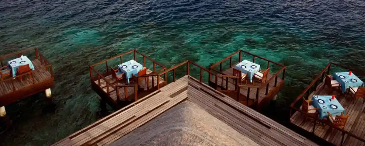 Coco Bodu Hithi, dining, North Malé Atoll, Maldives, Aqua restaurant signature dining pods: multiple individual mini-deck pods extending over the reef-shallow lagoon, each containing a single dining table set with light-blue tablecloth, timber-rail surround, the reef pattern visible beneath in the deep teal-cyan water (the property's most-photographed dining configuration).