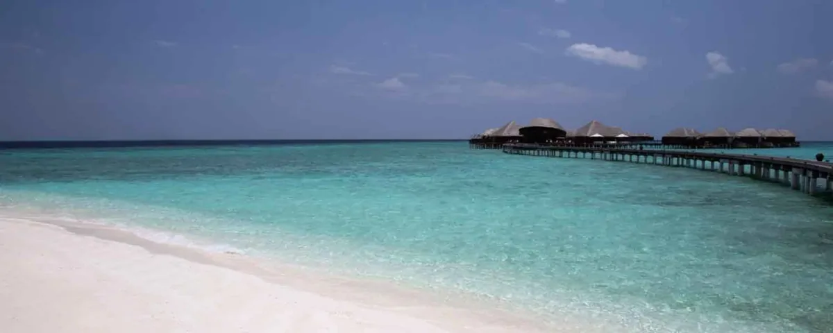 Coco Bodu Hithi, beach, North Malé Atoll, Maldives, Beach view from the lagoon side: white-sand foreground extending into the vibrant turquoise lagoon, the over-water villa row with thatched roofs visible at the right side extending out into the deep cobalt ocean, blue sky with a few clouds.
