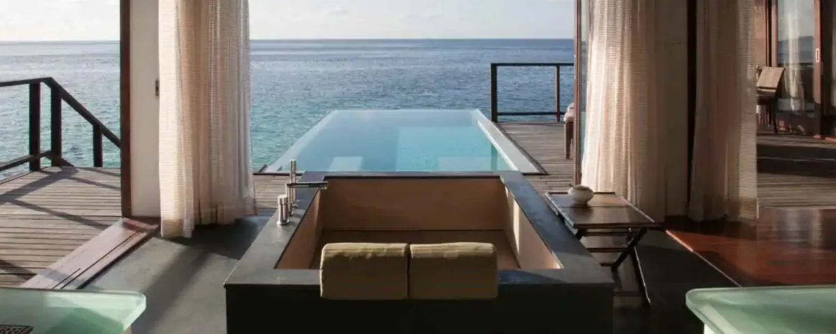 Coco Bodu Hithi, villa overwater, North Malé Atoll, Maldives, Coco Residence bath-pool axis: looking out through the villa interior across a deep timber bathtub (with twin head-cushion details), beyond it an infinity-edge plunge pool extending toward the ocean horizon, gauzy white curtains framing the view at the sides, a glass-top side table and partial timber deck visible.