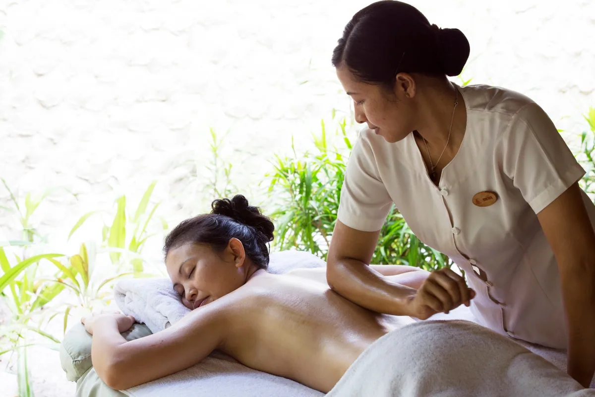 Coco Palm Dhuni Kolhu, spa, Baa Atoll, Maldives, Coco Spa treatment room, the therapist in cream Coco-branded uniform working a back massage beside the textured coral-stucco wall, the green planting and bamboo-veil providing the open-pavilion light.