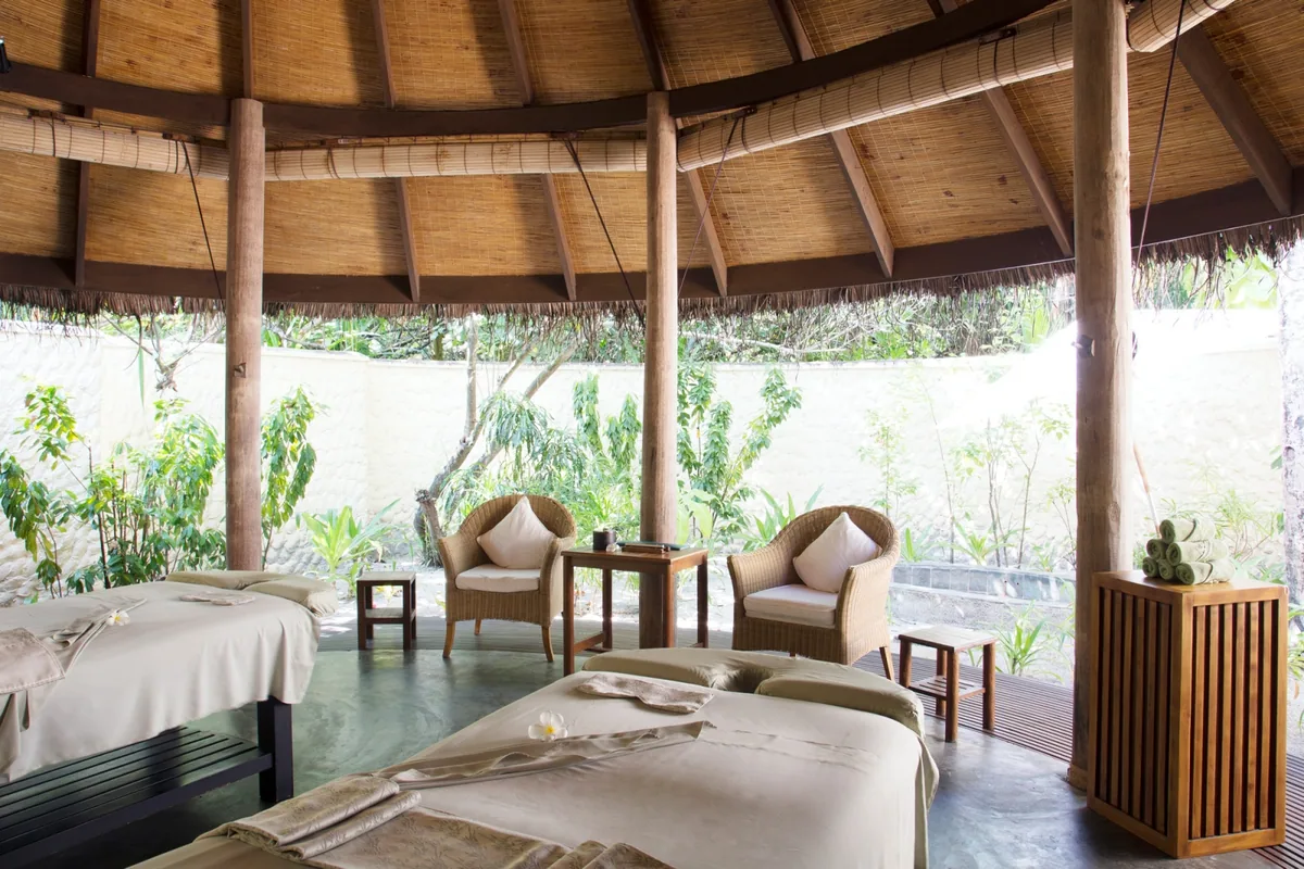 Coco Palm Dhuni Kolhu, spa, Baa Atoll, Maldives, Coco Spa twin treatment pavilion under the soaring conical thatched roof, the paired teak-frame massage beds with cream linens and plumeria-flower topping, woven wicker armchairs and polished concrete floor opening to the textured-stucco wall and tropical garden.