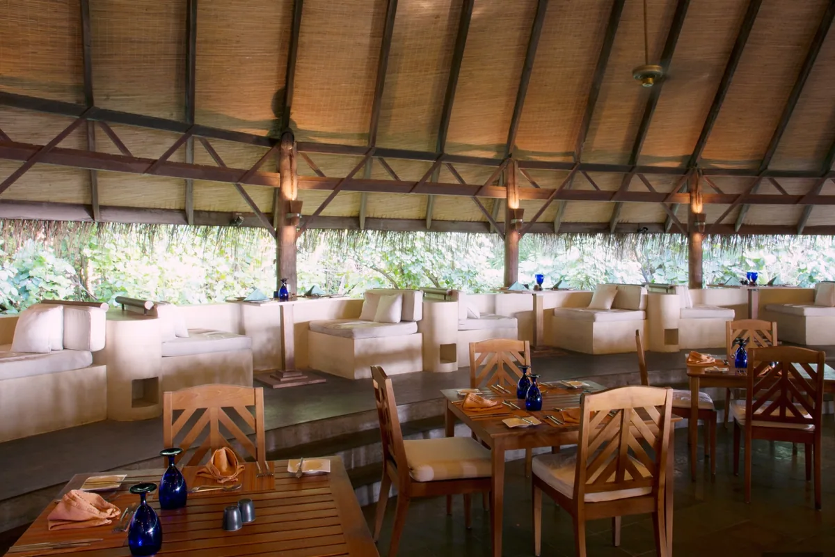 Coco Palm Dhuni Kolhu, dining, Baa Atoll, Maldives, Cowrie restaurant interior, the open-sided thatched-pavilion principal venue with the cobalt-blue glassware on raw-wood tables, woven palm-frond ceiling lattice and cream-stucco low banquette seating opening to the tropical garden.