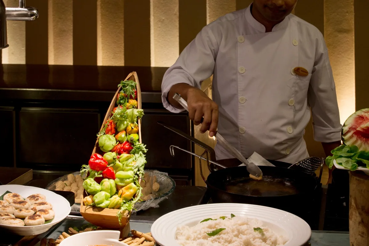 Coco Palm Dhuni Kolhu, dining, Baa Atoll, Maldives, Cowrie live-cooking station, the chef in pressed white jacket tossing the wok over the fire with the dhoni-shaped wooden vessel of red and green peppers, jasmine rice and watermelon arranged at the buffet edge.
