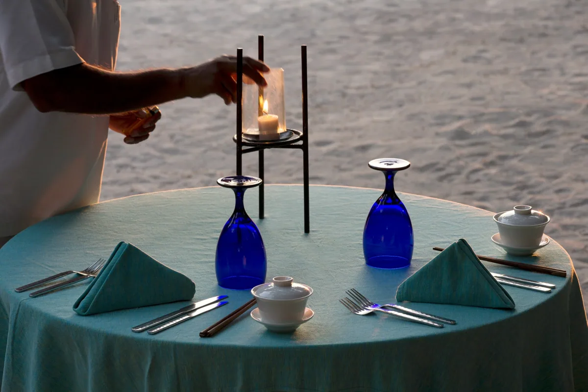 Coco Palm Dhuni Kolhu, dining, Baa Atoll, Maldives, Private beach dinner setting at sunset, the teal linen-covered round table with twin cobalt-blue Coco-signature glasses inverted on saucers, white porcelain rice bowls with lids and a glass-pillar candle lantern being lit by the white-jacketed server.