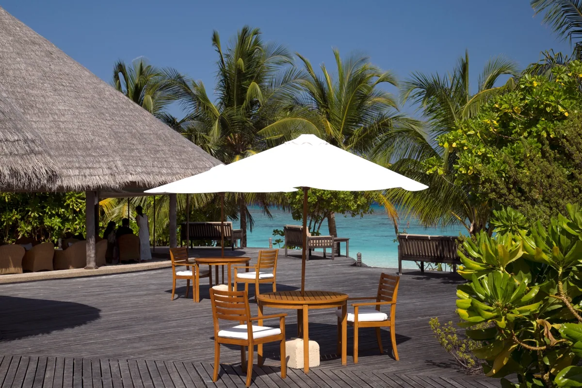 Coco Palm Dhuni Kolhu, dining, Baa Atoll, Maldives, Conch Bar deck, the open thatched-eave bar pavilion with the cream-canvas umbrella over the four-seat teak table on weathered hardwood decking, palm trees framing the turquoise lagoon directly beyond.