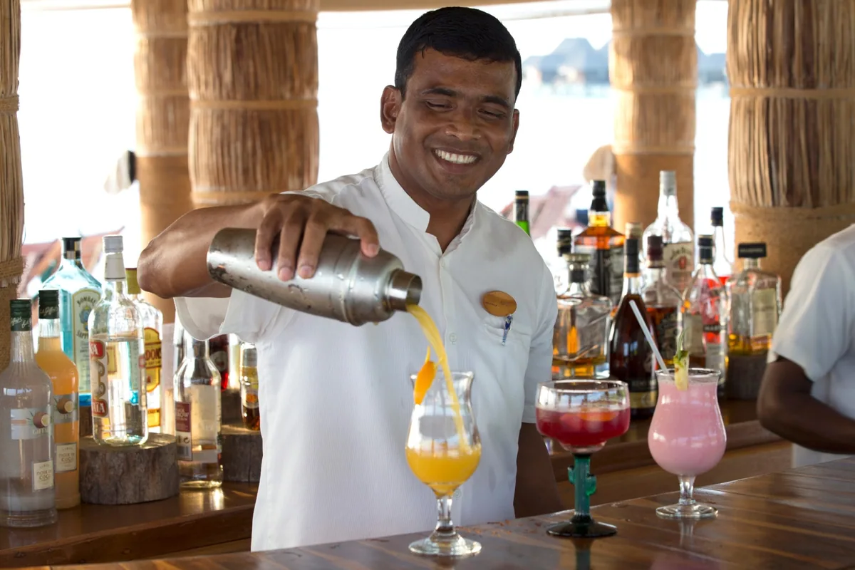 Coco Palm Dhuni Kolhu, dining, Baa Atoll, Maldives, Beach Bar bartender pouring a tropical cocktail from the shaker into three highball glasses, the woven palm-thatch column posts framing the back-bar bottle wall and the lagoon visible through the open eave.