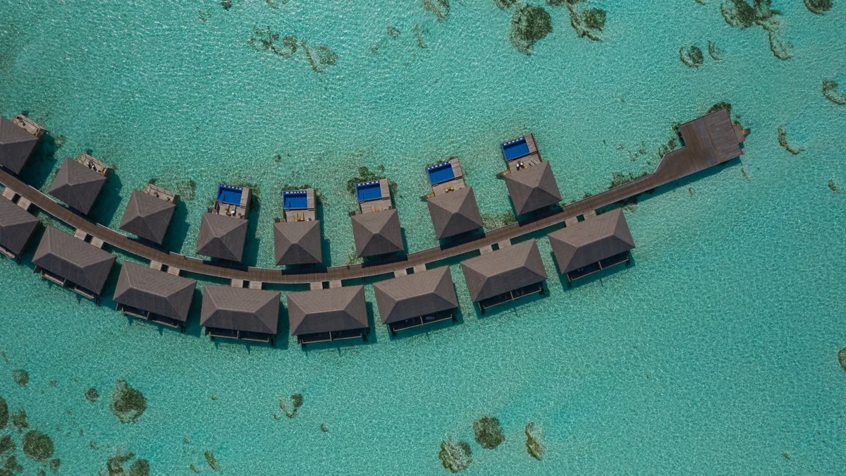 Cocoon Maldives, aerial, Lhaviyani Atoll, Maldives, Top-down aerial of the curved overwater-villa row with private plunge pools on the deck of each villa and reef patches scattered through the turquoise lagoon.