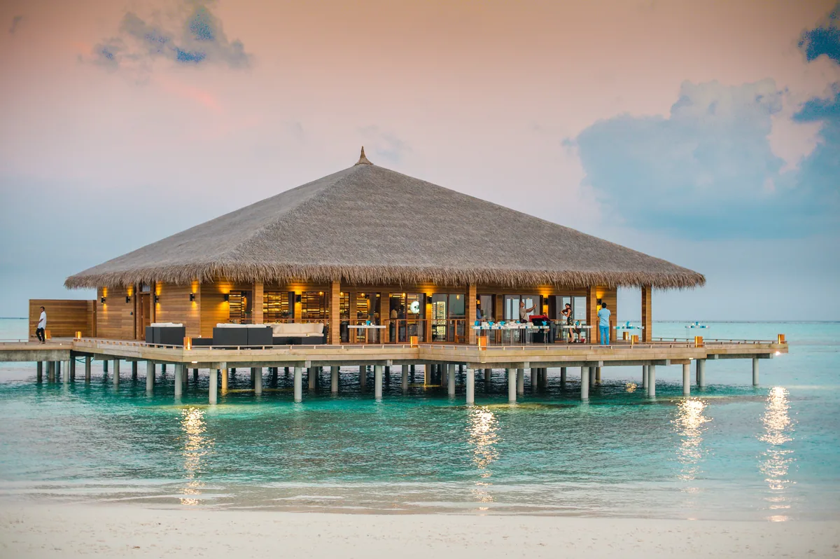 Cocoon Maldives, dining, Lhaviyani Atoll, Maldives, Manta principal all-day pavilion as a thatched pyramid pavilion sitting over the lagoon at sunset, glowing warm from within against the dusk sky.