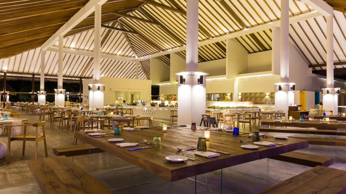 Cocoon Maldives, dining, Lhaviyani Atoll, Maldives, Octopus restaurant interior with white pillars under an A-frame open-beam roof, long wooden communal tables with benches, and lantern candles set for service.