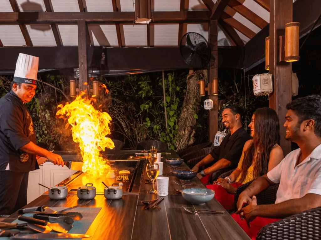 Cocoon Maldives, dining, Lhaviyani Atoll, Maldives, Japanese teppanyaki counter with the chef working a live flame, paper-lantern lighting overhead, and three guests seated at the counter.