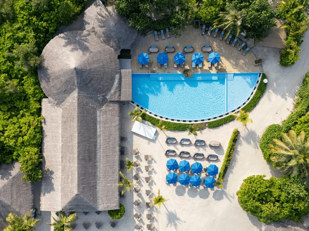 Cocoon Maldives, dining, Lhaviyani Atoll, Maldives, Top-down aerial of the Loabi Loabi pool bar showing the thatched pavilion adjacent to the curved infinity pool, blue umbrellas, and a ring of beach cabanas.