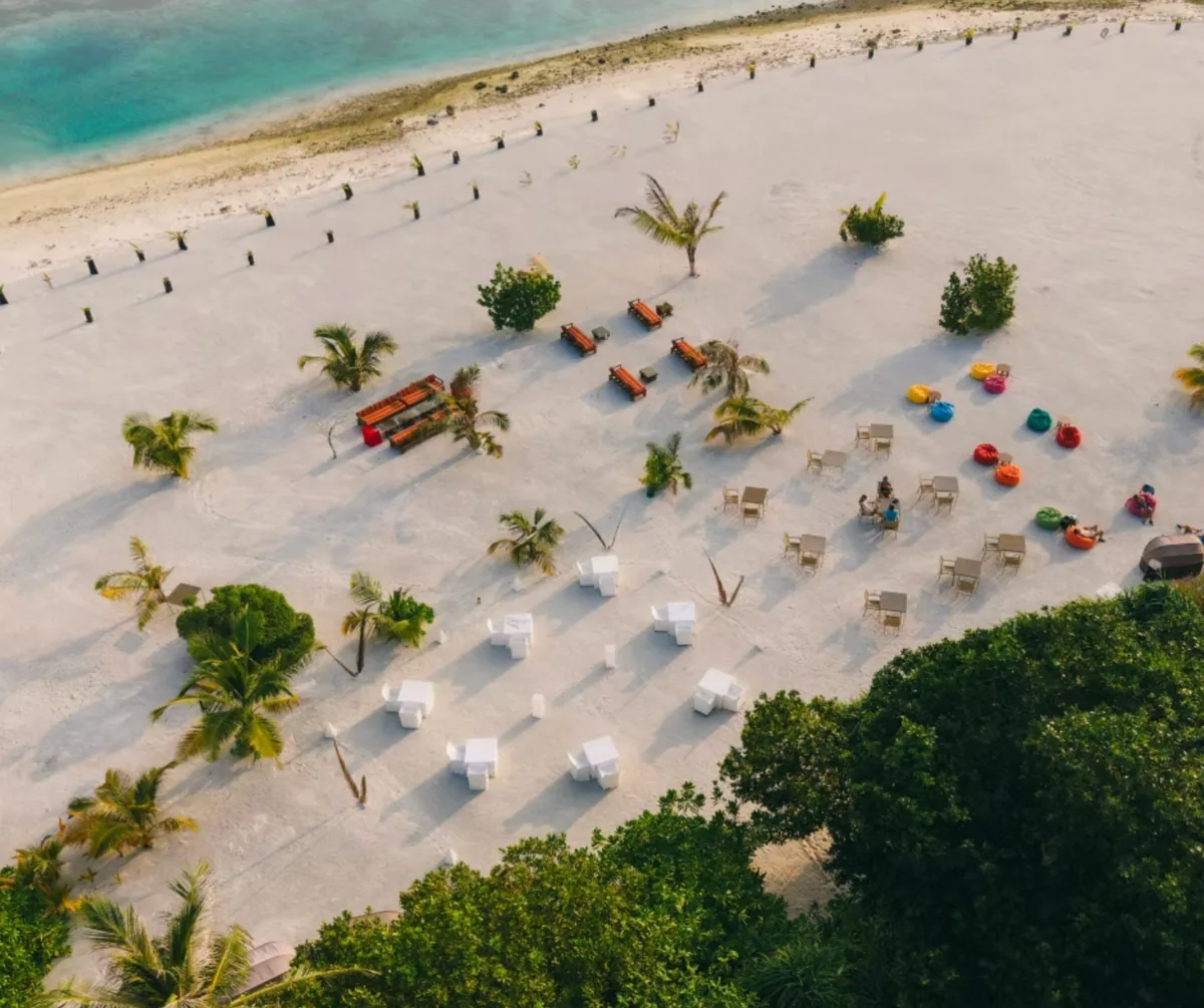 Cocoon Maldives, dining, Lhaviyani Atoll, Maldives, Aerial of the Kurum beach venue with colourful bean bag lounges, low orange-and-red benches, and scattered white lantern tables set across the sand.