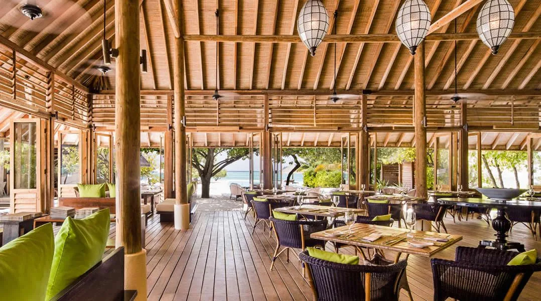COMO Cocoa Island, dining, South Malé Atoll, Maldives, Ufaa restaurant pavilion interior, open timber-pavilion architecture with vaulted slatted ceiling and exposed posts, lime-green cushioned banquette on the left, rattan dining chairs around timber tables, woven-shade pendant lanterns above and the lagoon visible past the open eaves at the rear.