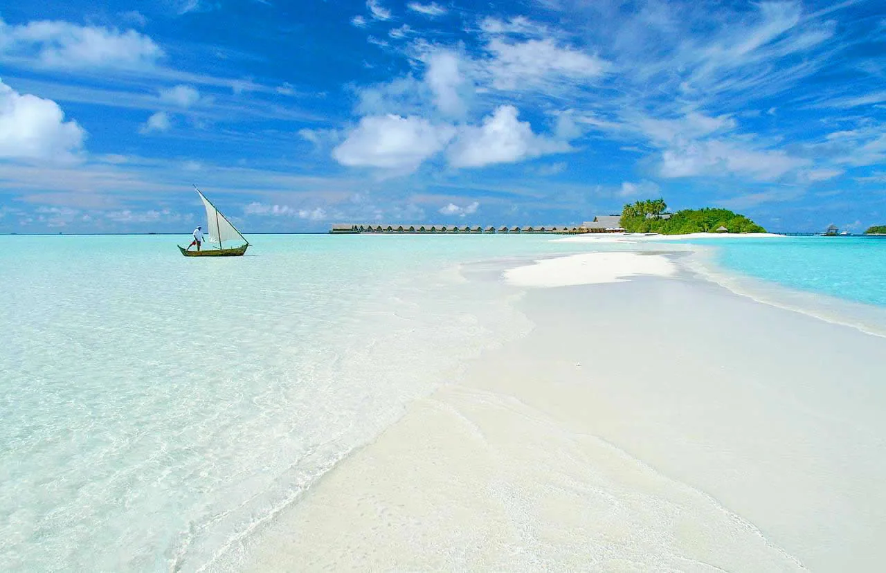 COMO Cocoa Island, setting, South Malé Atoll, Maldives, Sandbank shot with traditional dhoni sailboat in the foreground, a single figure rigging the white sail in the shallow water and the main island with its dhoni-shaped over-water villa row visible across the channel.
