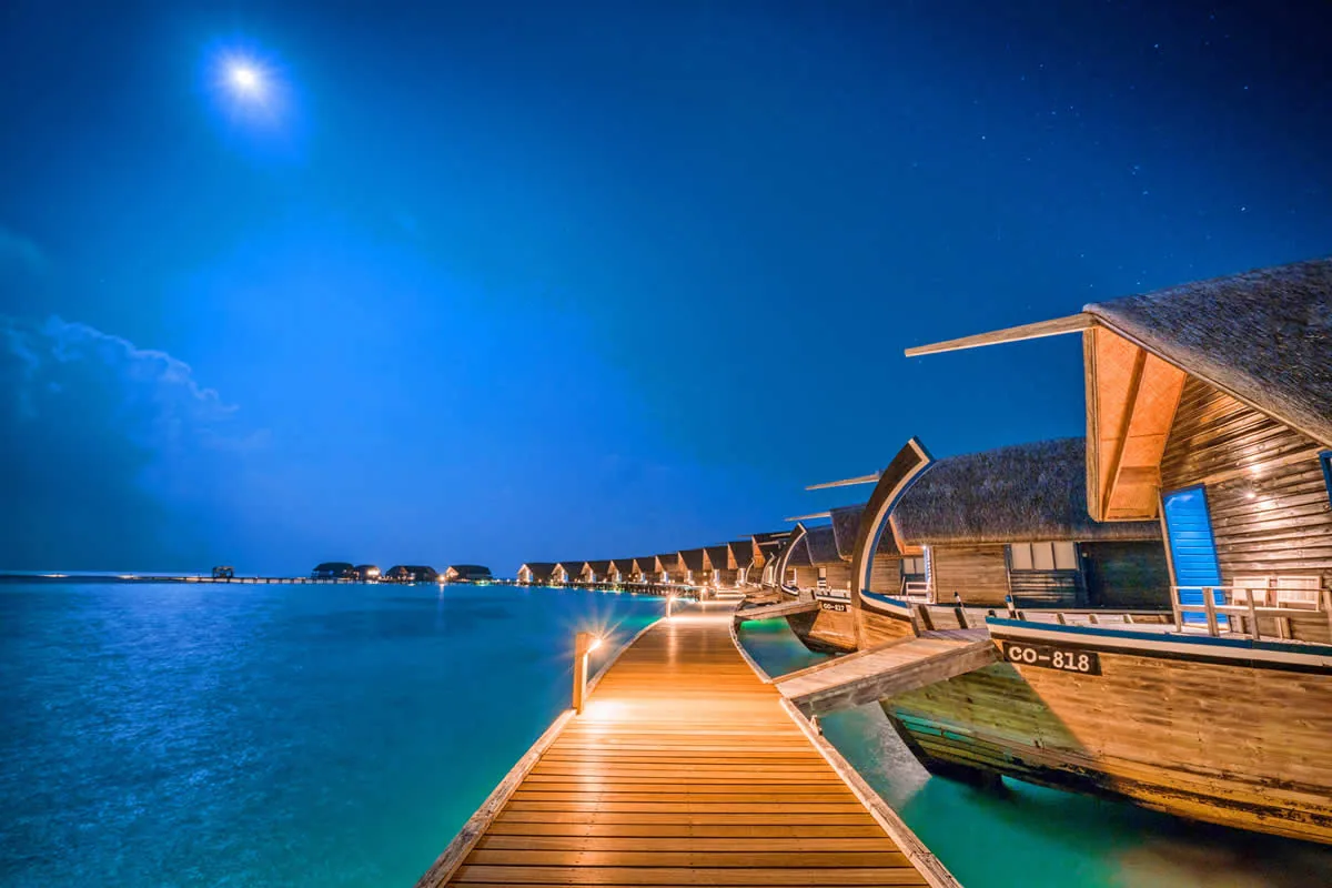 COMO Cocoa Island, setting, South Malé Atoll, Maldives, Daytime aerial of the villa arc, the curved over-water cluster reading clearly above the turquoise reef shelf with the connecting walking jetty looping back to the palm-canopy main island and the boat jetty exiting left into the deeper-water channel.