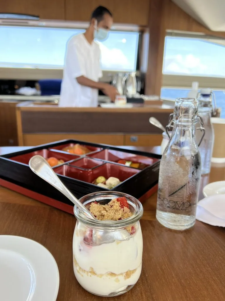 COMO Cocoa Island, arrival, South Malé Atoll, Maldives, Aboard the COMO Cocoa Island yacht transfer, a parfait jar of yogurt with granola and berries on a blonde-timber bento board, a COMO Cocoa Island branded glass bottle of still water on the right, a uniformed steward preparing service in the open salon behind.