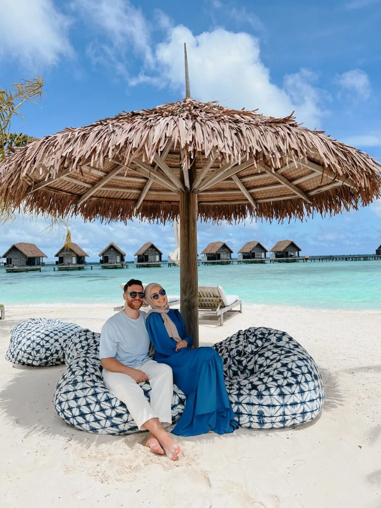 COMO Cocoa Island, setting, South Malé Atoll, Maldives, Beach lounge moment under a palm-frond parasol, two guests seated on indigo-shibori bean-bag loungers facing the lagoon, the curved dhoni-shaped over-water villa row reading in profile on the horizon line behind.
