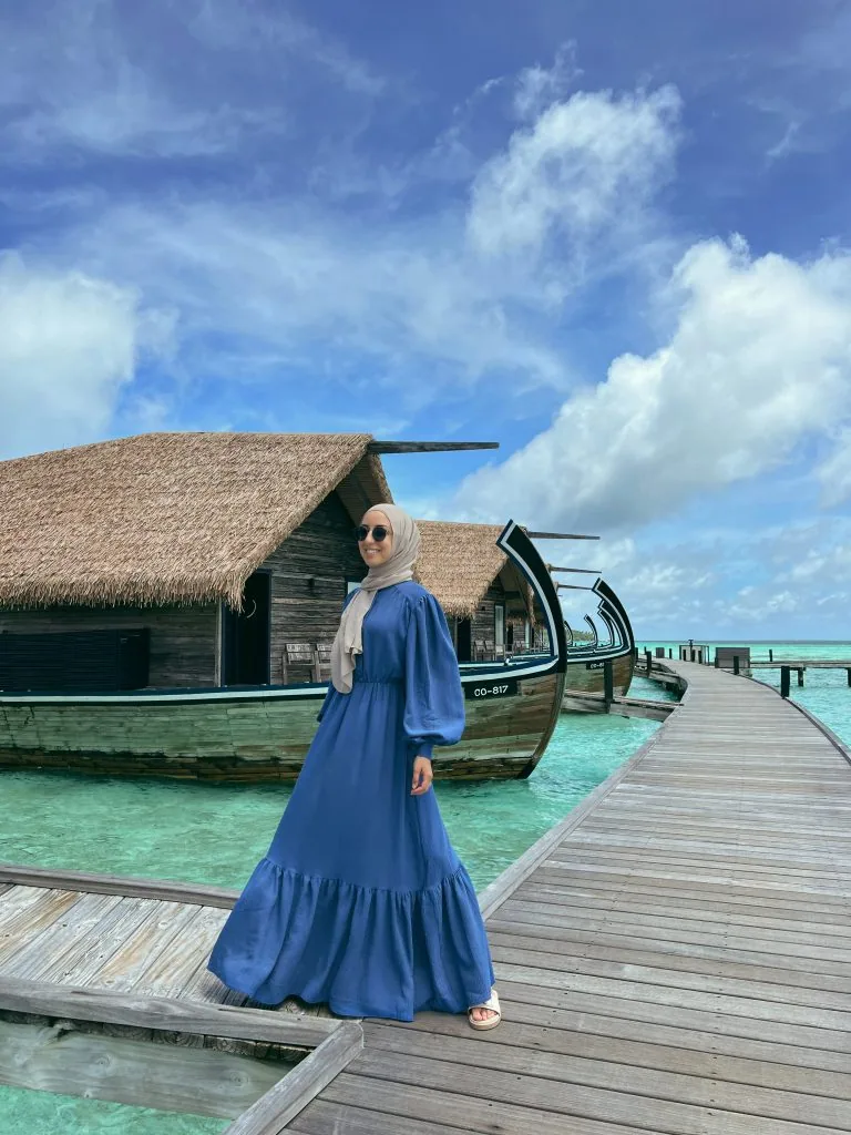 COMO Cocoa Island, villa overwater, South Malé Atoll, Maldives, Boardwalk-side perspective of a dhoni villa, the curved timber hull profile visible at right with villa number CO-817 on its bow and the thatched gable roof above, a guest in a blue dress on the timber walkway for scale, the empty boardwalk continuing right out to the further villa cluster.