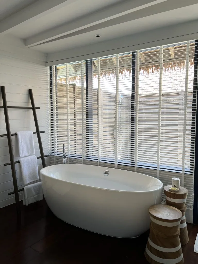 COMO Cocoa Island, villa overwater, South Malé Atoll, Maldives, Dhoni villa bathroom, signature COMO white-on-white palette: oval freestanding tub on dark hardwood floor, white-shutter Venetian blinds across full-height windows revealing the thatched eave of the adjacent villa, dark-timber ladder towel rail, carved-stripe stools beside the tub.