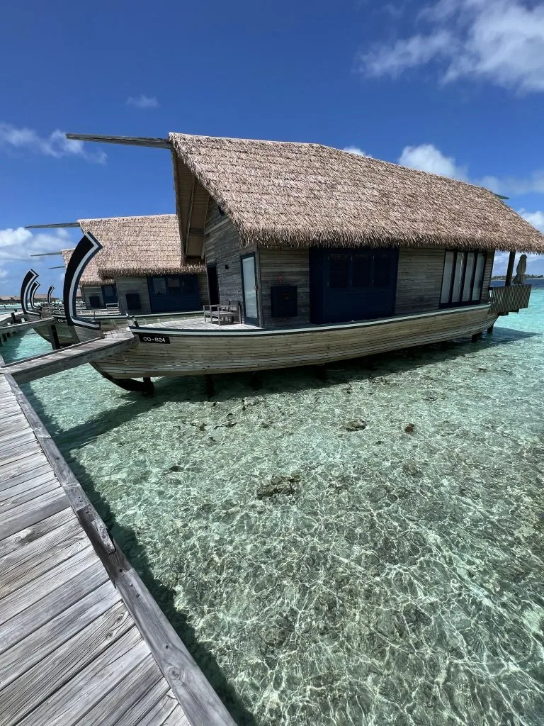 COMO Cocoa Island, villa overwater, South Malé Atoll, Maldives, Single dhoni-shaped villa close-up (CO-RO4), the curved timber hull profile under a steeply pitched thatched roof, dark shutters and a small private rear deck reaching back to the boardwalk.
