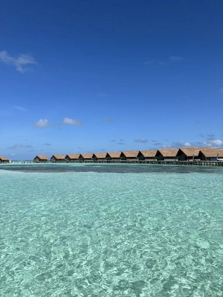 COMO Cocoa Island, villa overwater, South Malé Atoll, Maldives, Full villa row from the lagoon, the line of thatched pitched roofs reading in profile from the open water, the boardwalk just visible behind connecting each villa back to the main island.