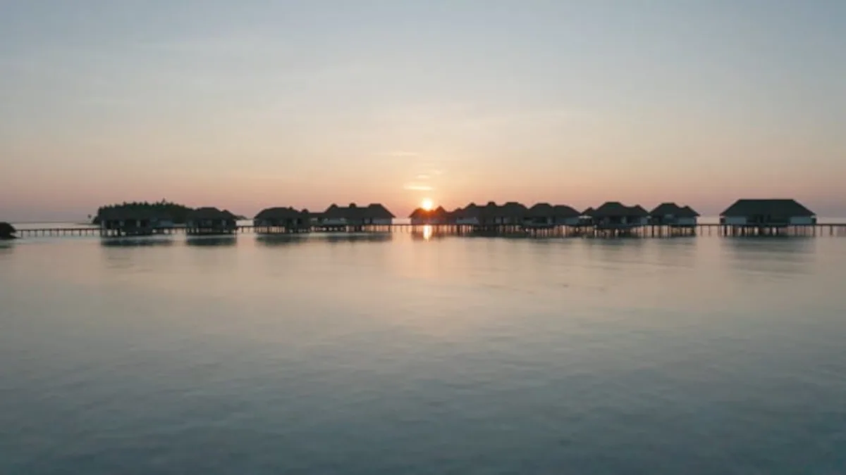 COMO Maalifushi, setting, Thaa Atoll, Maldives, The signature over-water villa cluster at sunset, silhouettes of the thatched-roof villa row across the calm mirror lagoon with the sun touching the horizon between the villas.