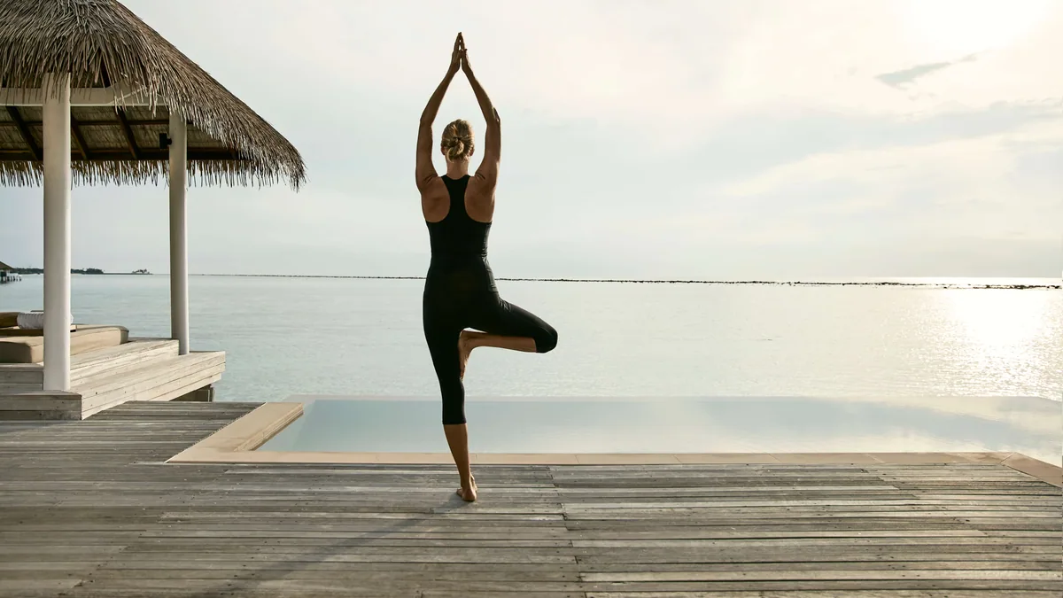COMO Maalifushi, spa, Thaa Atoll, Maldives, COMO Shambhala wellness pavilion, a guest in tree-pose yoga on the timber deck, thatched pavilion at the left edge, an infinity-edge plunge pool at the deck's outer line, mirror lagoon stretching to the horizon.