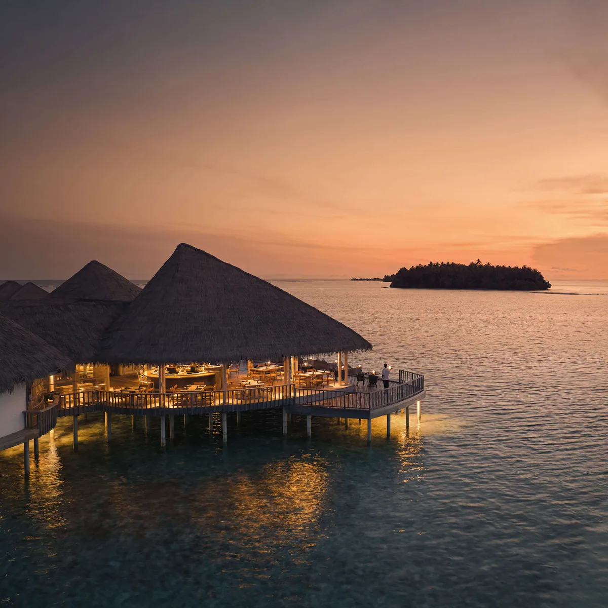 COMO Maalifushi, dining, Thaa Atoll, Maldives, Tai over-water restaurant at sunset, a thatched conical-roof pavilion lit from within over the lagoon, two figures on the deck looking out toward a distant island silhouette, orange sky reflecting on the calm lagoon below the pillars.