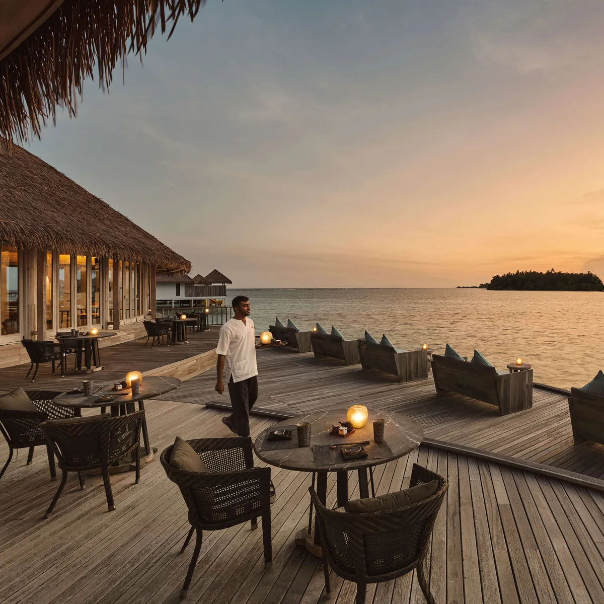 COMO Maalifushi, dining, Thaa Atoll, Maldives, Over-water dining deck at golden hour, a server walking between rounds of wicker dining tables and chairs with hurricane lanterns, the thatched-roof restaurant lit from within at the far edge, soft sunset light over the calm lagoon.