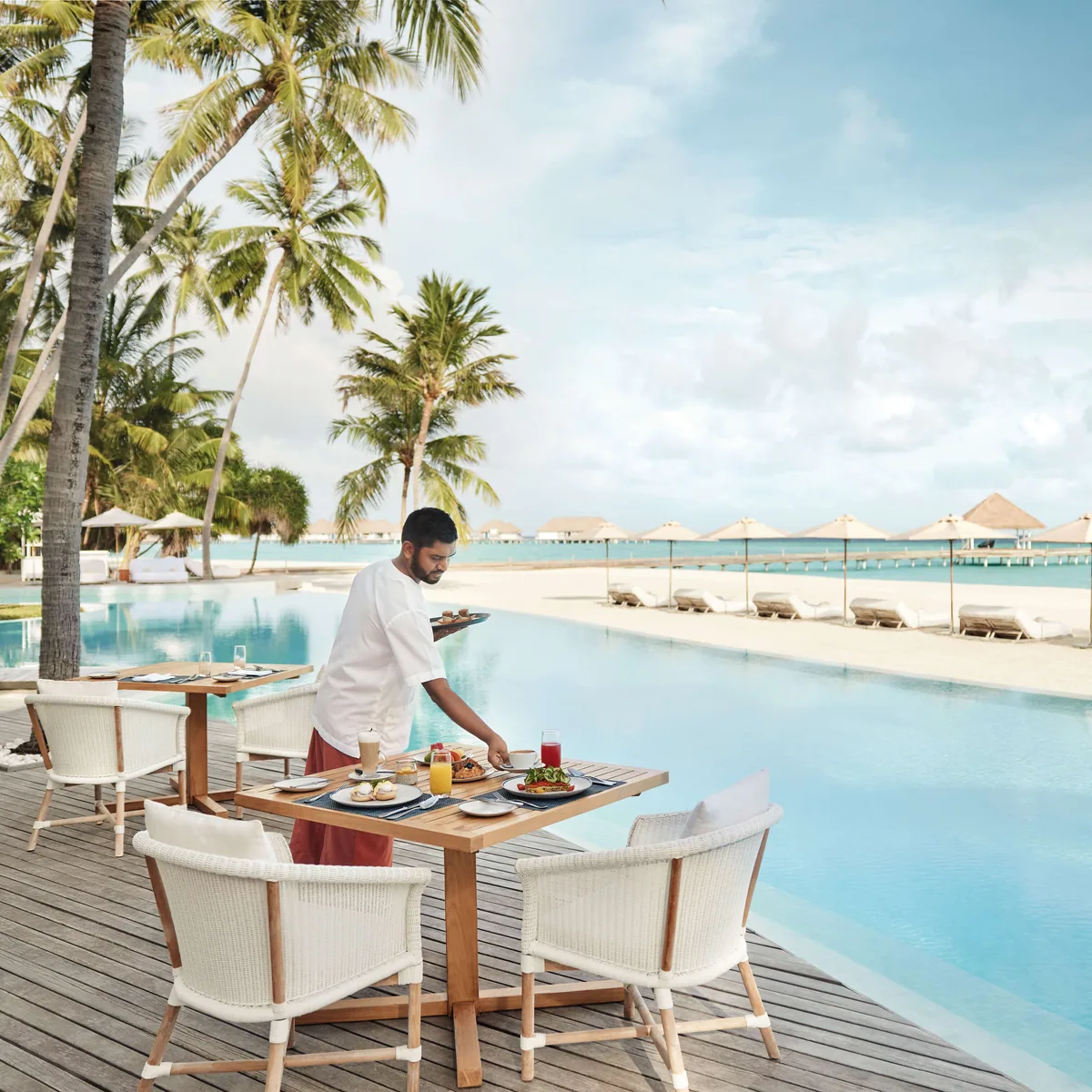 COMO Maalifushi, dining, Thaa Atoll, Maldives, Poolside restaurant at breakfast, a server delivering a tray to a wicker-chair table beside the long main pool, palms framing the left edge, thatched-roof cabanas and the white-sand beach with the over-water villa cluster visible at the back.