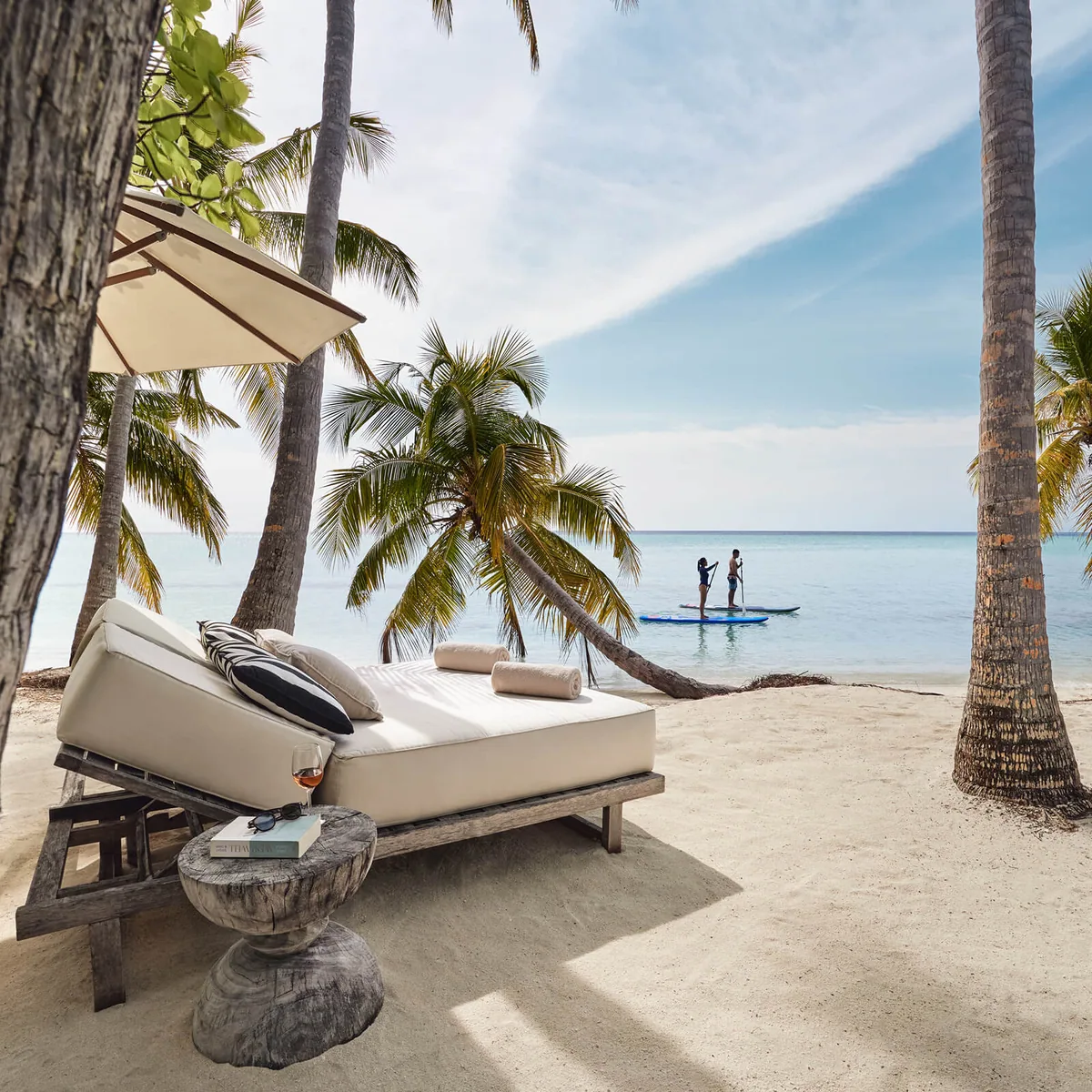 COMO Maalifushi, setting, Thaa Atoll, Maldives, Beach daybed lifestyle moment, a large white-cushioned double daybed under a canvas shade strung between palm trunks, a paddleboard couple in the lagoon beyond, white-sand beach in the foreground.
