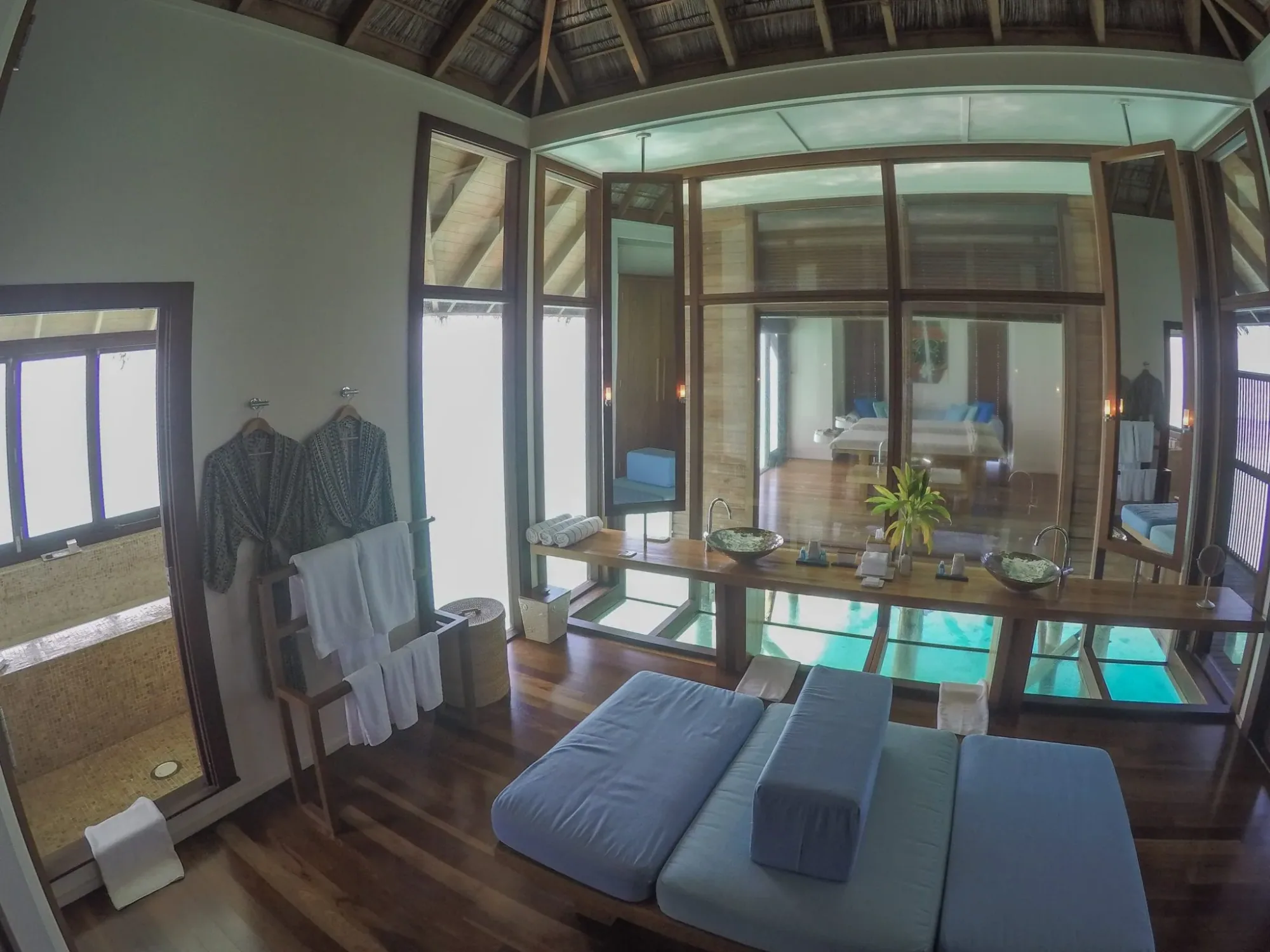 Conrad Maldives Rangali Island, villa overwater, Alif Dhaal (South Ari) Atoll, Maldives, Water Villa bathroom with the in-floor coral-viewing glass panel, twin spa massage bed, hanging robes and an outdoor shower beyond the timber wall, an early example of the glass-floor signature later adopted across the property.