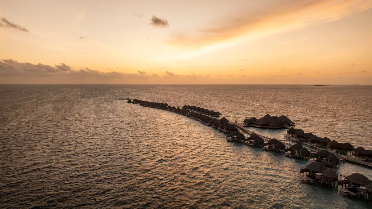 Constance Halaveli, aerial, Alif Alif Atoll (North Ari), Maldives, Iconic dhoni-hull water-villa arc aerial at sunset, the two curving overwater villa rows tracing the dhoni-boat outline against the golden lagoon, the high-pitched pavilion architecture along the main island catching the late light. The property's signature visual identity.