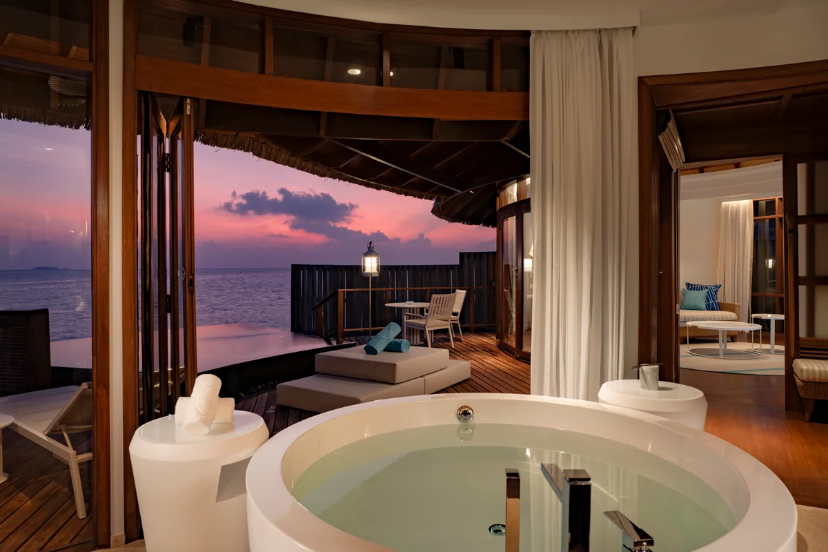 Constance Halaveli, villa overwater, Alif Alif Atoll (North Ari), Maldives, Water Villa with Pool private deck at twilight, built-in turquoise-tile lap pool extending the length of the deck with a glass-walled overwater shower visible beyond, the pavilion-style thatched-roof villa structure at the rear, deep teal lagoon catching the dusk light.