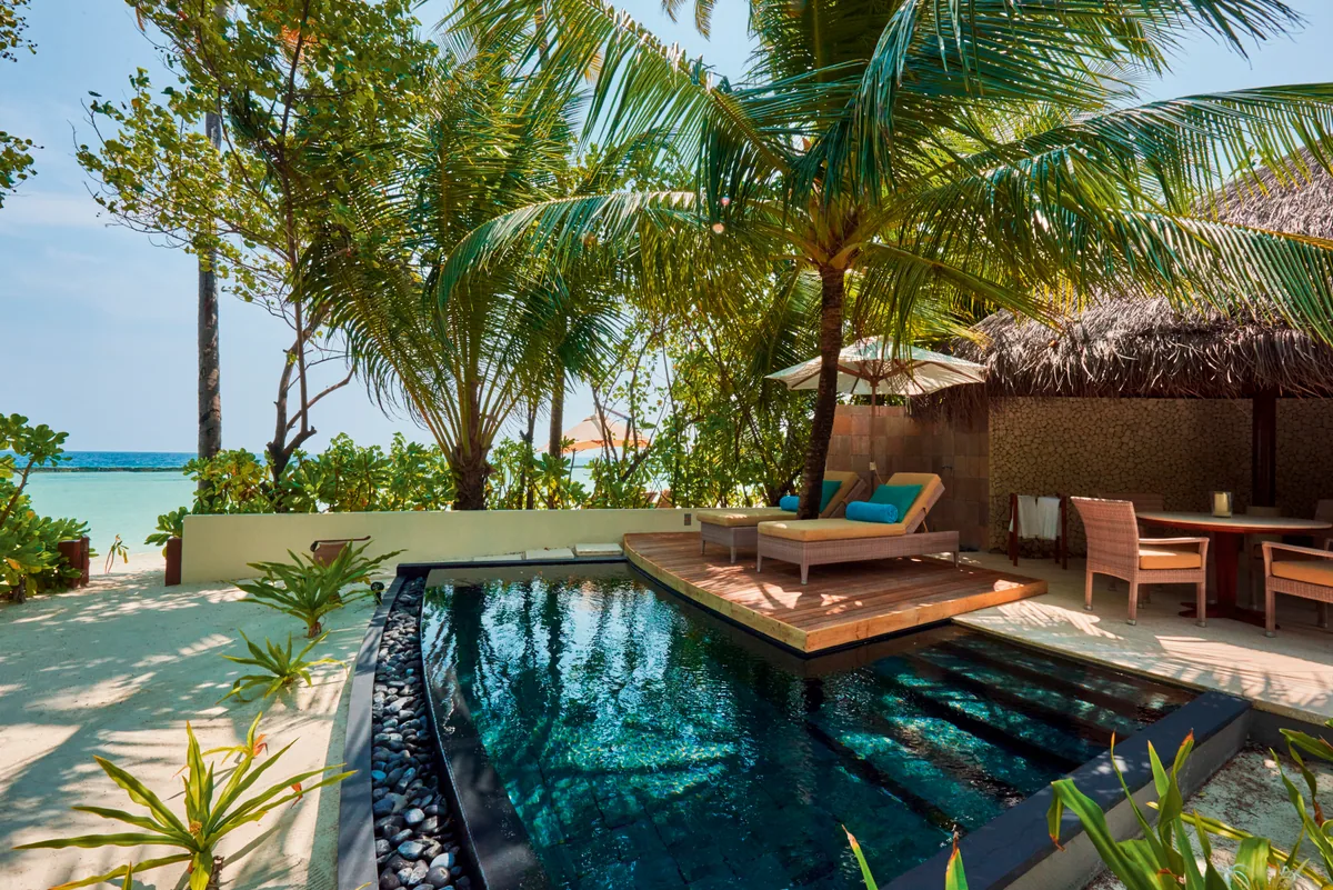 Constance Halaveli, villa beach, Alif Alif Atoll (North Ari), Maldives, Beach Villa with Pool, perimeter sand line with thatched cabana over the dark-stone-framed plunge pool, the pavilion-vernacular interior visible through the open doors, Mauritian-influenced detailing in the wall treatments.