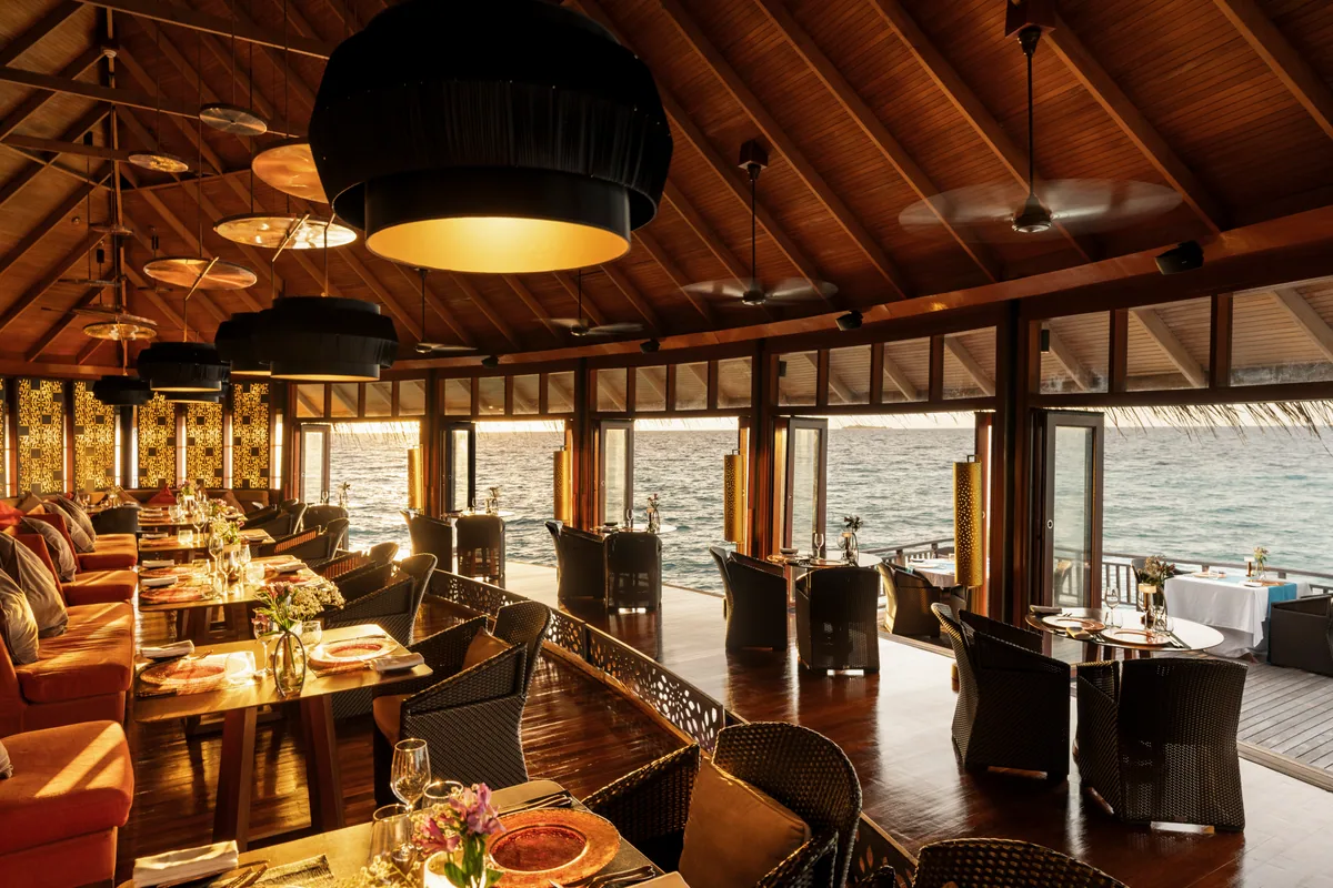 Constance Halaveli, dining, Alif Alif Atoll (North Ari), Maldives, Jing overwater restaurant interior at sunset, high-pitched timber-rafter ceiling with black drum-pendant lights, warm-lit dining room with woven-back chairs around tables set in copper and brass place settings, terracotta banquette running the left wall, lattice Asian-pattern screens on the columns, ocean visible through three sides of full-height windows.