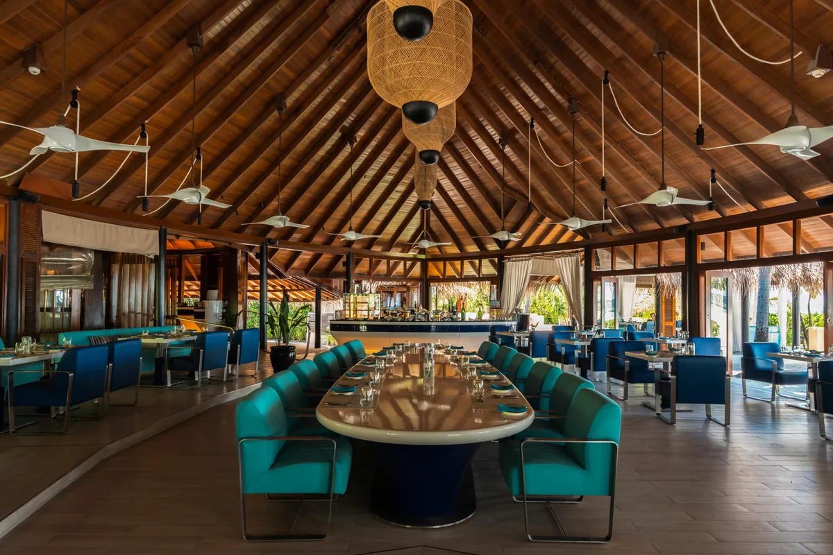 Constance Halaveli, dining, Alif Alif Atoll (North Ari), Maldives, Jahaz Restaurant pavilion interior, high-pitched timber-rafter ceiling with woven-raffia round pendant lights, central long dark-navy oval communal table surrounded by turquoise and blue woven-mesh chairs (Mauritian color signature), dark wood floor and the bar reading at the rear.