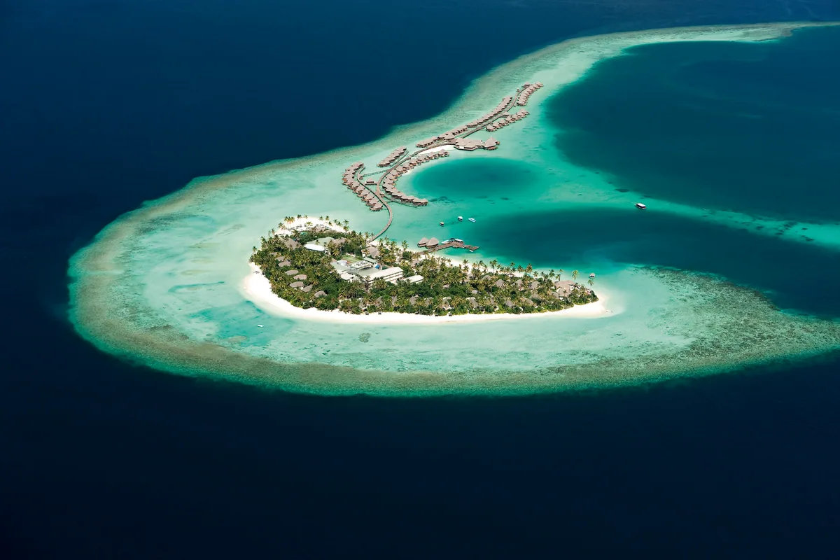 Constance Halaveli, setting, Alif Alif Atoll (North Ari), Maldives, Daylight aerial of the dhoni-shape island layout, palm-covered main island with thatched-roof pavilions in the centre, overwater villas extending in two curving rows from each end forming the Maldivian-dhoni outline, the reef-protected turquoise lagoon encircling and the deep open ocean beyond.