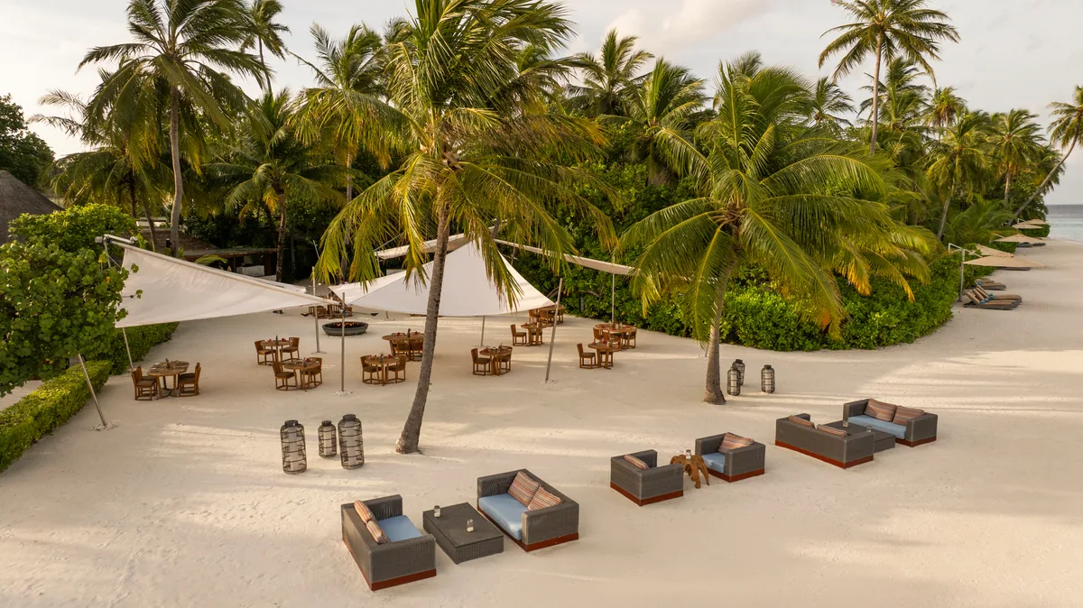Constance Halaveli, setting, Alif Alif Atoll (North Ari), Maldives, Beach lounge setting with white-canvas shade sails strung between palms, low woven sofas with blue cushions in clusters on the white sand, lantern arrangements on the deck, palm canopy framing the lagoon view beyond. The Mauritian-pavilion-style outdoor public realm in daily-use mode.