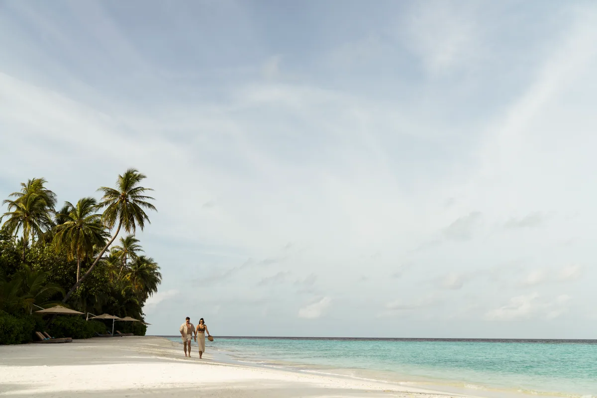 Constance Halaveli, lifestyle, Alif Alif Atoll (North Ari), Maldives, Couple walking the Halaveli beach in soft midday light, palm canopy framing the left side of the frame, the property's sun-loungers under conical canvas umbrellas visible at the tree line, the turquoise lagoon stretching to the horizon.
