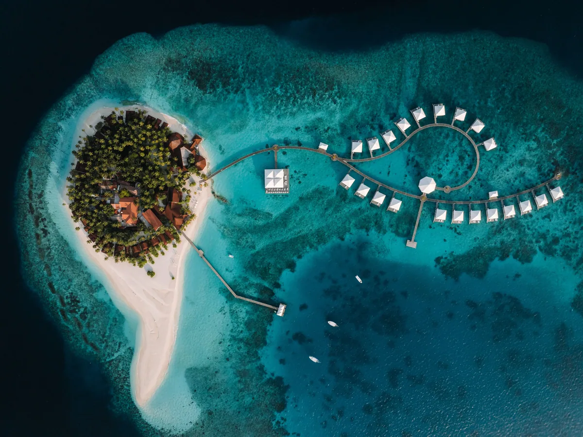 Diamonds Thudufushi Beach & Water Villas, aerial, Alif Dhaal (South Ari) Atoll, Maldives, G-loop overwater walkway aerial, the curved boardwalk threading the orange-roof overwater pavilion into the lagoon, the property's sandbank-extension main island in the frame and the encircling reef on the central South Ari Atoll edge.