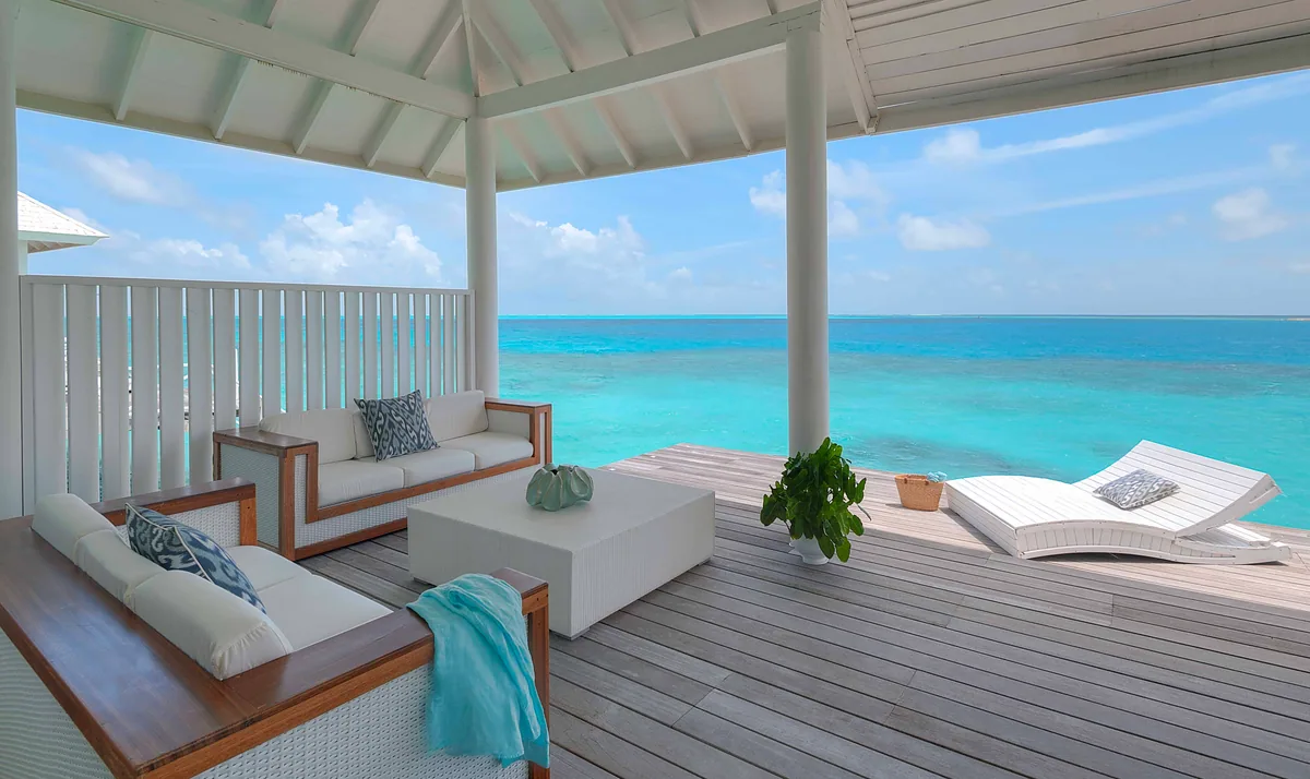 Diamonds Thudufushi Beach & Water Villas, villa overwater, Alif Dhaal (South Ari) Atoll, Maldives, Over Water Villa Suite private deck, peaked-rafter ceiling visible through the open sliding doors, the wood deck running over the turquoise lagoon, twin loungers under the pavilion overhang.