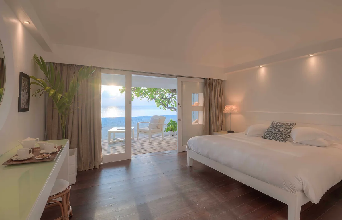 Diamonds Thudufushi Beach & Water Villas, villa beach, Alif Dhaal (South Ari) Atoll, Maldives, Standard Beach Villa sunset deck, king bed visible through the open doors with white linen and pillows, the wooden deck extending onto the sand at sunset, palm canopy framing the view.