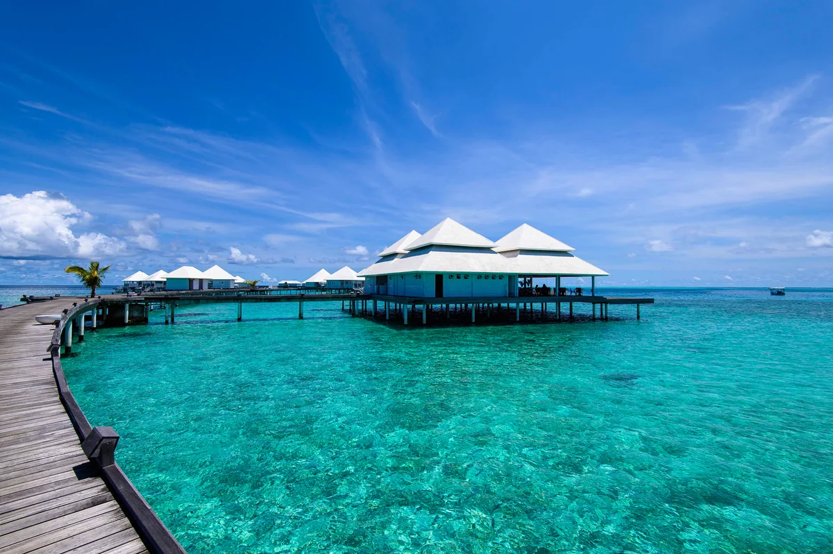 Diamonds Thudufushi Beach & Water Villas, dining, Alif Dhaal (South Ari) Atoll, Maldives, Aqua Over Water Restaurant exterior, peaked tent-style white roof pavilion on stilts at the end of a curving wooden boardwalk, crystal-clear turquoise lagoon with reef texture visible beneath, palm at the left.