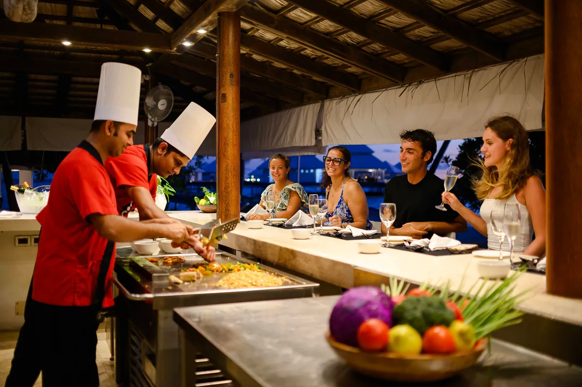 Diamonds Thudufushi Beach & Water Villas, dining, Alif Dhaal (South Ari) Atoll, Maldives, Teppanyaki counter at night, two chefs in red-and-black uniforms grilling at the metal counter while four guests sit at the counter watching with wine glasses, thatched-roof pavilion overhead, palm canopy and main island visible behind.