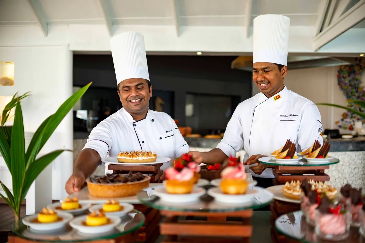 Diamonds Thudufushi Beach & Water Villas, dining, Alif Dhaal (South Ari) Atoll, Maldives, Two pastry chefs in white toques arranging the tiered dessert spread (lemon tarts, mousse, fruit-topped cakes) on wooden display stands, sunlit indoor restaurant background, plants framing the right edge. The Italian-management lineage food-side cadence.