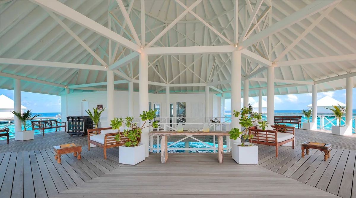 Diamonds Thudufushi Beach & Water Villas, spa, Alif Dhaal (South Ari) Atoll, Maldives, Centro Serena Spa pavilion interior, white-painted timber-frame pitched roof with mint-green ceiling underside, woven raffia pendant lampshades, light-grey hardwood floor, wooden bench seats flanking a central console with potted plants, the deck and lagoon visible through arched openings on either side.