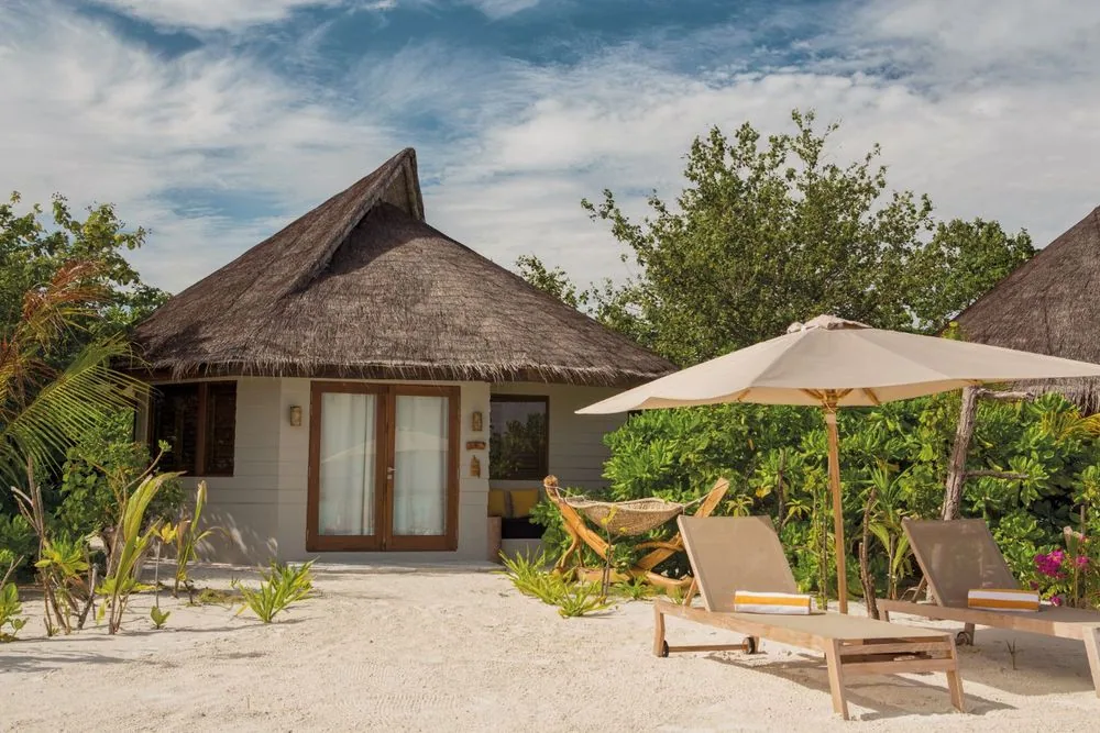 DRIFT Thelu Veliga Retreat, villa beach, Alif Dhaal (South Ari) Atoll, Maldives, Beach Villa exterior with conical thatched roof, single-pavilion white-walled bungalow tucked into the palm canopy, sun-loungers under a wood-frame canvas umbrella and a striped hammock slung between rough-timber posts on the white sand.