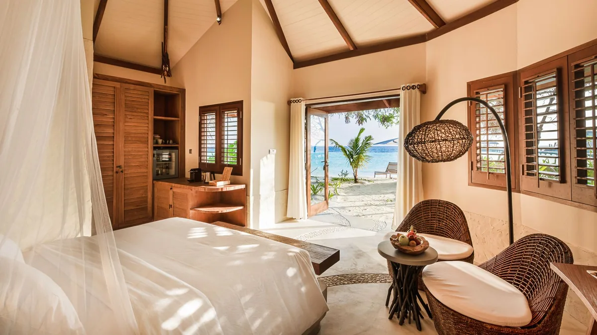 DRIFT Thelu Veliga Retreat, villa beach, Alif Dhaal (South Ari) Atoll, Maldives, Beach Villa interior, cathedral pitched-rafter ceiling with timber beams, king bed under a draped mosquito net canopy, woven-rattan basket pendant lamp, woven egg chair and side table, plantation-shutter walls, open door framing the beach loungers and lagoon view.